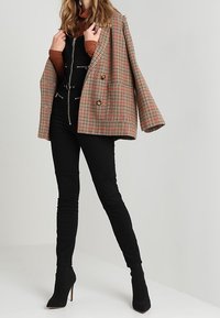 Blazer de cuadros de corte cruzado en beige, rojo y marrón; superpuesto sobre un mono negro con cremallera; combinado con pantalones negros ajustados y botas de tobillo.
