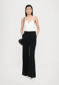 Un débardeur blanc froncé avec de fines bretelles, associé à un pantalon noir taille haute à jambes larges. Le mannequin tient un sac à main noir et porte des talons légers.