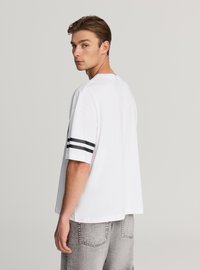 Biała T-shirt z krótkim rękawem, czarne paski na rękawach, luźny krój, wykonana z bawełny, gładka tekstura, warstwowy detal przy dolnym brzegu.