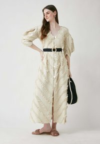 Beige midi-mekko, jossa on teksturoitu sahalaitakuosi, suuret napit ja vyötetty mustalla vyöllä. Yhdistetty ruskeisiin matalapohjaisiin sandaaleihin ja mustaan laukkuun.