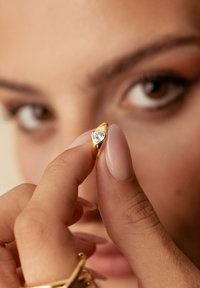Anello d'oro a forma di cuore, con un diamante trasparente al centro. Mano con unghie di colore chiaro che tiene l'anello, con gioielli visibili.