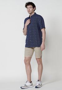 Camicia a maniche corte blu navy con un motivo testurizzato, abbinata a pantaloni corti beige arrotolati e sneakers bianche con dettagli grigi e blu navy.