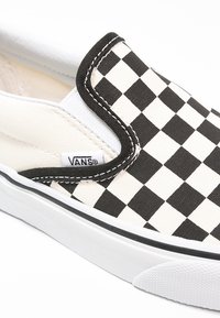 Scarpa slip-on in tela a scacchi bianco e nero con suola in gomma bianca e cuciture nere, dotata di un piccolo etichetta con il logo Vans sul lato.