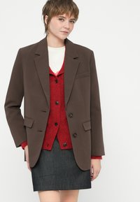 Sacou brun, croit special, cu rever în formă de U, două buzunare frontale și trei butoane. Purtați peste un cardigan texturat roșu și o fustă din denim.