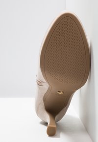 Chaussure à talon haut beige présentée talon relevé contre une surface blanche, montrant la semelle texturée et le talon aigu fin.