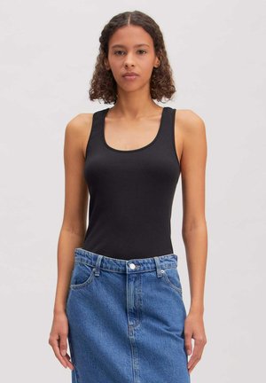 Jonge vrouw die een zwart tanktop draagt, ingestopt in een hoog getailleerde blauwe spijkerrok, staand tegen een effen lichte achtergrond.