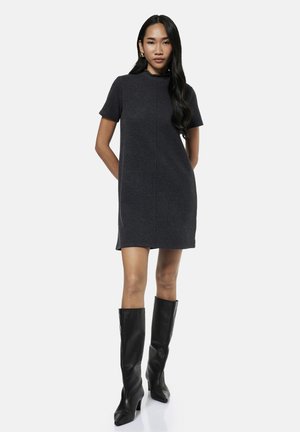 Femme debout, les mains dans le dos, portant une robe gris foncé à manches courtes et des bottes noires montantes sur un fond uni.