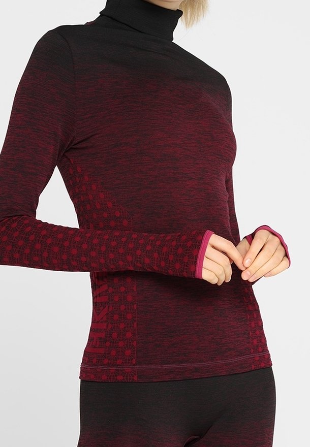 Kvinde iført en bourgogne- og sort langærmet tætsiddende top med tommelhul og matchende leggings, poserende med hænderne nær taljen mod en ensfarvet baggrund.