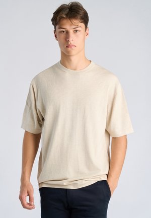 Jonge man met kort bruin haar, gekleed in een loszittend beige overhemd met korte mouwen en een donkere broek, staand tegen een lichtgrijze achtergrond.