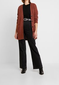 Brauner gestrickter Cardigan mit Knöpfen, weiten Ärmeln und einer Vorderseite mit Tasche, kombiniert mit hoch taillierter schwarzer Denimhose und schwarzen Stiefeletten.