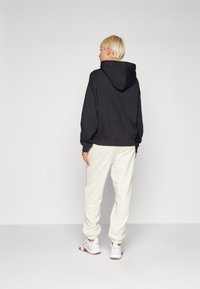 Čierna mikina s kapucňou vo voľnejšom strihu, skombinovaná so svetlo béžovými teplákmi. Biely športový obuv s hnedými podrážkami dokončuje tento outfit.