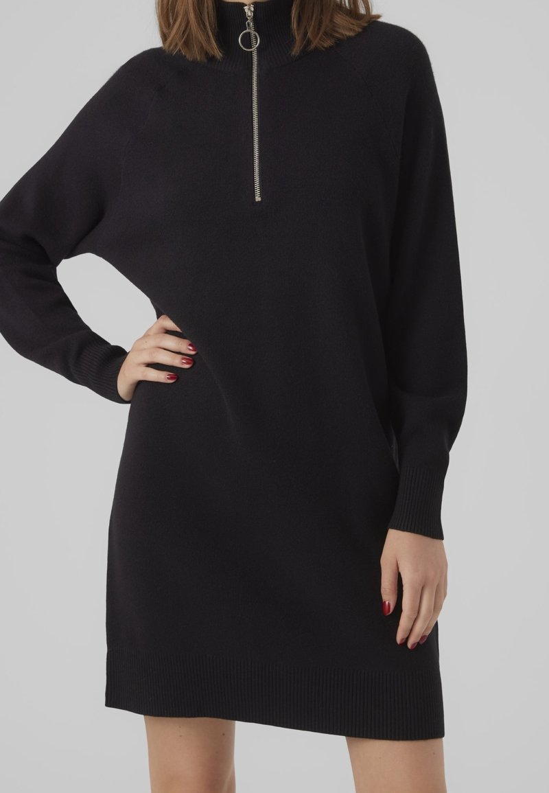 Femme portant une robe en maille noire à manches longues avec une fermeture éclair devant et des poignets côtelés, posant avec une main sur la hanche.