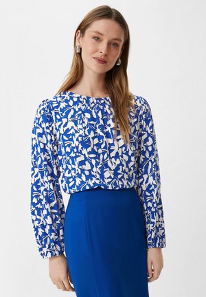 Donna con capelli lunghi che indossa una blusa floreale blu e bianca con maniche lunghe e una gonna blu a tinta unita, posando su uno sfondo neutro.