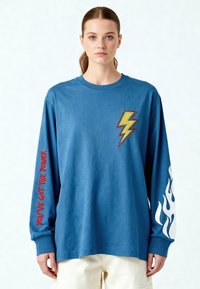 Jeune femme portant une chemise bleue à manches longues avec un éclair jaune sur la poitrine, un texte rouge « VOUS AVEZ LE POUVOIR » sur la manche gauche, et un dessin de flamme blanche sur la manche droite.