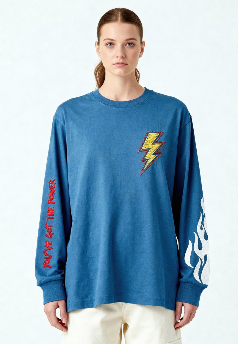 Jeune femme portant une chemise bleue à manches longues avec un éclair jaune sur la poitrine, un texte rouge « VOUS AVEZ LE POUVOIR » sur la manche gauche, et un dessin de flamme blanche sur la manche droite.