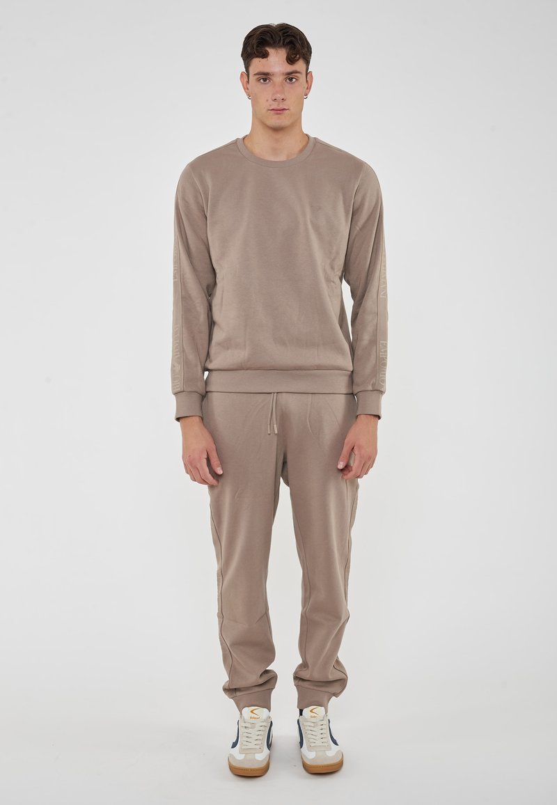 Felpa beige e pantaloni jogger abbinati in tessuto liscio, con polsini a coste e vita con cordino, indossati con sneakers bianche e blu.