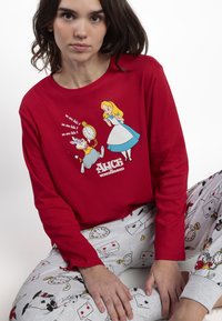 Mujer vestida con una camiseta de manga larga roja con estampado de Alicia en el País de las Maravillas y pantalones grises decorados con naipes y relojes de bolsillo.