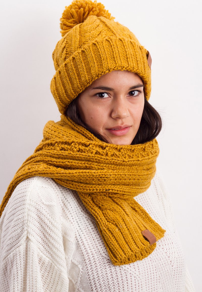 Yellow knitted beanie with a pom-pom and matching thick scarf. Both feature textured patterns and leather accent tags, set against a light background.