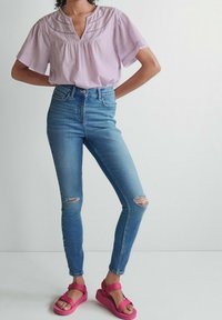 Lys lilla kortærmet top med V-hals og dekorativ syning, parret med slidte blå skinny jeans og lys pink sandaler.
