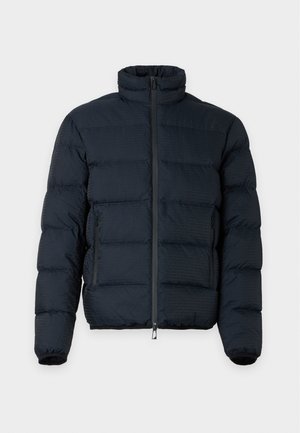 Nuori mies seisoo päällään musta toppatakki, tummat revityt farkut ja ruskeat lenkkarit, yksiväristä valkoista taustaa vasten.
