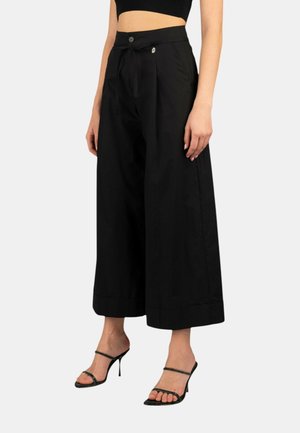 Pantaloni neri a gamba larga realizzati in materiale leggero, con vita alta, chiusura con bottone e dettagli sul fondo. Abbinati a tacchi neri con lacci.