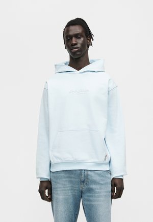 Μαύρος άνδρας με πλεξούδες, φορώντας ένα ανοιχτό μπλε φούτερ και ξεθωριασμένο μπλε τζην, στέκεται μπροστά σε απλό λευκό φόντο.