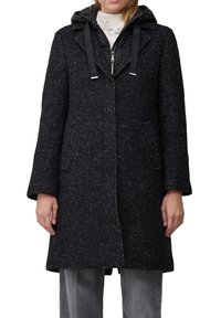 Vrouw in een knielange donker gesprenkelde jas over een zwarte rits-hoodie en grijze broek, staande tegen een witte achtergrond.
