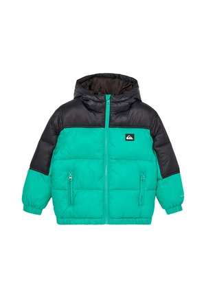 Enfant portant une doudoune turquoise et noire, un bonnet noir, des lunettes de soleil rouges réfléchissantes et un pantalon bleu foncé, debout devant un fond uni.