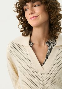 Maglione lavorato a maglia color crema con un design a lavorazione aperta, colletto a V e una camicia con stampa floreale a contrasto sotto. Tessuto morbido, vestibilità casual.