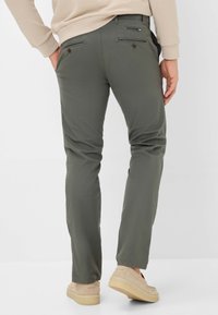 Olive green pants with a slim fit, featuring two back pockets with button closures, paired with beige shoes. Soft, textured fabric.