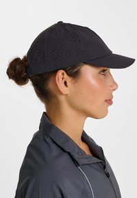 Schwarze Mütze mit durchlöchertem Design zur Belüftung, ausgestattet mit einer gebogenen Krempe und verstellbarem Riemen hinten. Graue Jacke mit Reißverschluss.