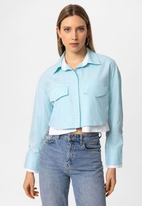 Lyseblå cropped skjorte med lange ærmer og to forlommer, lagd over en hvid undertrøje, parret med højtaljede blå jeans.