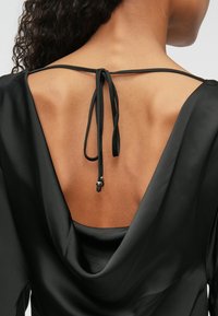 Melnā blūze ar kapuci un dziļu V formas aizmuguri, kurai ir saišu detaļa ar metālisku akcentu un gludu, satīna līdzīgu tekstūru.