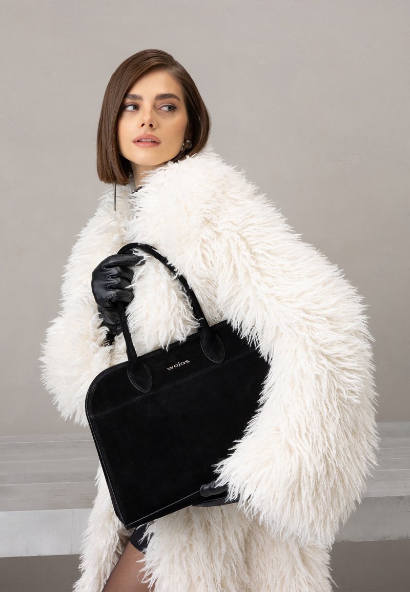 Černá semišová kabelka s krátkými uchy a detaily s logem, držená v ruce s rukavicí. Modelka má na sobě huňatý bílý kabát, který přidává kontrast textur.