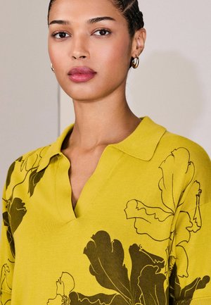 Žluté pletené polo tričko s výstřihem do V a krátkými rukávy, zdobené tmavě hnědými květinovými vzory. Hladká textura, minimální množství kovových detailů.