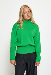 Roheline kudumiga kaeluse-sweater pikkade varrukatega, millel on istuv taljeosa ja pehme tekstuur. Kombineeritud mustade pükstega.