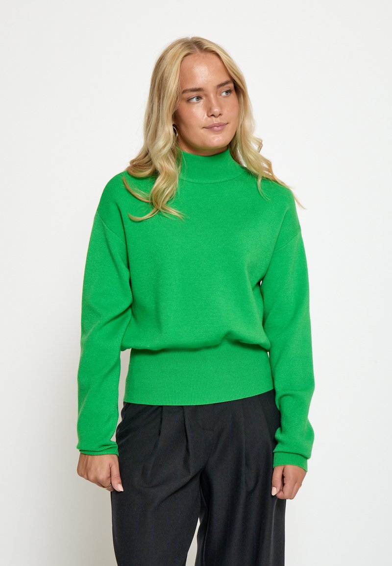 Roheline kudumiga kaeluse-sweater pikkade varrukatega, millel on istuv taljeosa ja pehme tekstuur. Kombineeritud mustade pükstega.