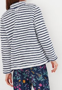 Kobieta ubrana w biało-granatowy paskowany, puszysty żakiet i granatowe, kwiatowe spodnie, widoczna z tyłu na tle gładkiej, białej ściany.