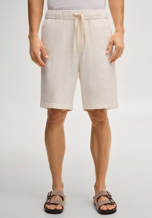 Mężczyzna noszący beżowe, teksturowane szorty z troczkiem, białą koszulę i brązowe sandały z klamrą, stojący na jednolitym tle.