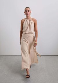 Beige, halterförmiges Oberteil aus texturiertem Stoff, kombiniert mit einem langen, schwingenden Rock mit seitlichem Bindeband und fransigem Saum. Schwarze Sandalen vervollständigen den Look.