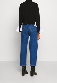 Personne portant un jean bleu droit, des bottines noires à talons blocs, et un pull noir avec des détails blancs aux poignets, debout de dos.