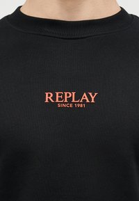 Μαύρο φούτερ με στρογγυλή λαιμόκοψη και κόκκινο κείμενο "REPLAY SINCE 1981" τοποθετημένο στο κέντρο του στήθους.