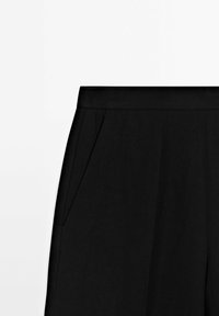 Pantalon noir sur mesure avec poches latérales et ceinture plate sur fond blanc uni.