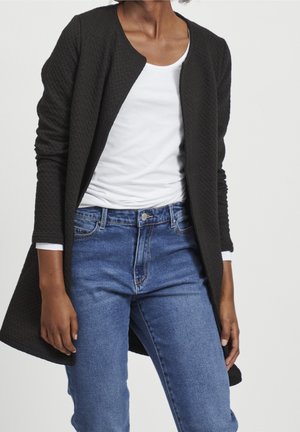 Femme portant un cardigan noir ouvert texturé, une chemise blanche à manches longues et un jean bleu sur fond uni.