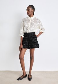 Blusa blanca de seda con estampado floral y botones en la parte delantera. Pantalones cortos negros de texturizados con volantes en capas y acentos plateados. Bailarinas negras.