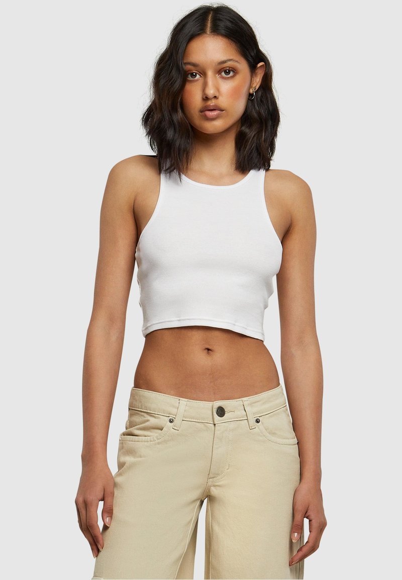 Femme aux cheveux foncés mi-longs porte un crop top blanc sans manches et un pantalon beige, debout devant un fond clair uni.