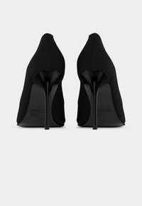 Μαύρα γυναικεία ψηλοτάκουνα γόβες στιλέτο με μυτερά δάχτυλα, ορατά από πίσω, που δείχνουν κομψά τακούνια και λείο ύφασμα.