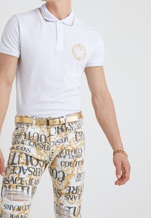 Mann trägt ein weißes Polohemd mit goldenen Rändern und Emblem, gemusterte weiße Jeans mit Text und goldenen Mustern, goldenen Gürtel und goldene Gliederarmband.