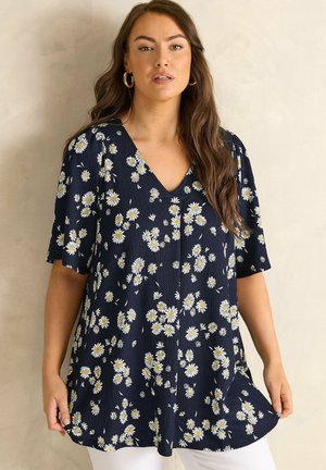 Frau trägt eine marineblaue V-Ausschnitt-Bluse mit weißen Gänseblümchenaufdrucken und kurzen Ärmeln, kombiniert mit weißen Hosen, steht vor einer schlichten Wand.