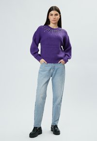 Lila gestrickter Pullover mit Verzierungen am Ausschnitt, kombiniert mit hellblauen Jeans und schwarzen Plateauschuhen.